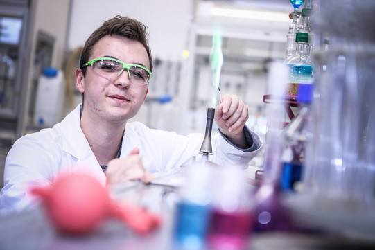 Männlicher Auszubildender mit Kittel und Schutzbrille im Chemielabor erzeugt eine grüne Flamme am Bunsenbrenner.